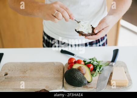 Junger Mann, der in der modernen weißen Küche Avocado-Toasts zubereitet und Käsecreme auf Vollkornbrot legt. Prozess der Herstellung von Toasts mit Avocado und Toma Stockfoto