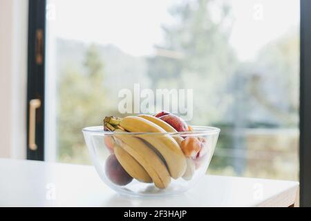 Reife tropische Früchte in stilvoller Glasschale auf Hintergrund großer Fenster in moderner Küche. Banane, Pfirsich, Aprikose auf der Arbeitsplatte. Sommervitamine. Heilen Stockfoto