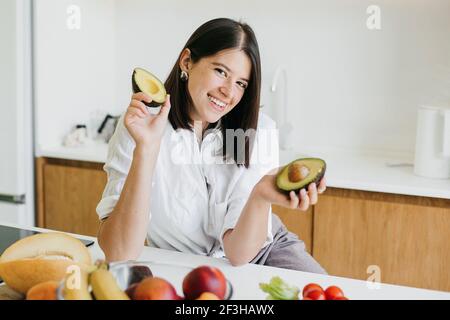 Junge glückliche Frau hält Hälften von reifen Avocado in den Händen und lächelt in der modernen weißen Küche mit frischem Obst und Gemüse. Gesunde Ernährung und Hom Stockfoto