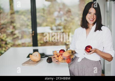 Junge glückliche Frau hält Schüssel mit sommerlichen Früchten an der weißen Arbeitsplatte mit Gemüse in der modernen weißen Küche. Gesunde Ernährung und Hausmannskost Konzept Stockfoto