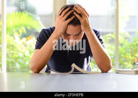 Junger Mann, der den Kopf mit den Händen hält, müde vom Lesen und Lernen Stockfoto