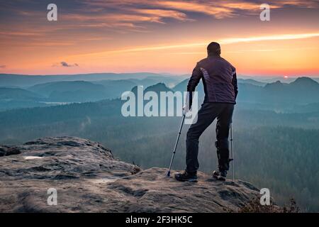 Behinderter Tourist mit Krücken. Rückansicht des verletzten Wanderers mit Unterarmstangen, die vor dem Hintergrund des Sonnenuntergangs am Berg laufen Stockfoto