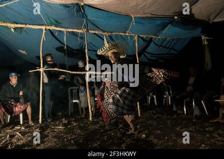 Ein älterer in traditioneller Kleidung tanzt vor dem Hintergrund von Männern, die während einer Gemeindeversammlung auf Rote Island, East Nusa Tenggara, Indonesien, Schlaginstrumente spielen. Stockfoto