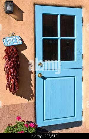 Eine turqupoise bemalte Holzstalltür in einer lehmziegelwand, mit einem Chili-Ristra und einem bemalten 'offenen' Schild, an der Canyon Road, Santa, Fe, New Mexico. Stockfoto