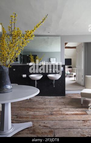 En-Suite Badezimmer mit zwei Wand aufgehängten Waschbecken auf schwarzem Mosaik Hintergrund mit Krug aus gelber Forsythia auf kreisförmigen grauen Tisch - Designer: Fox Linton Stockfoto