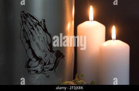 Eine Metallurne mit betenden Händen und brennenden Kerzen mit Asche von einem Toten bei einer Beerdigung. Trauriger trauernder Moment am Ende eines Lebens. Letzter Abschied Stockfoto