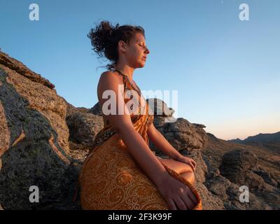 Seitenansicht des Mädchens mit einem seidenorangefarbenen Kleid, das im Sonnenuntergang auf einem Granitberg meditiert. Stockfoto