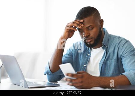 Besorgt schwarz Freiberufler Kerl Blick auf Smartphone während der Arbeit an Heimbüro Stockfoto