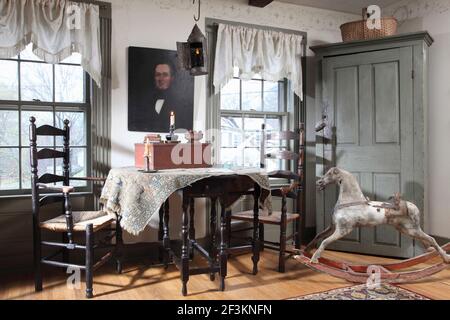 Ein informeller Raum mit Tisch, Stühlen und primitivem Eckschrank. Auch im Zimmer ist ein altes Schaukelpferd und ein Portrait über dem Tisch. Stockfoto