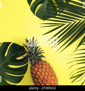 Draufsicht auf frische Ananas mit tropischer Palme und Monstera-Blättern auf gelbem Tischhintergrund. Stockfoto