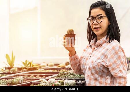 Authentische asiatische Frau lächelnd hält Kaktus in einem Kunststoff-Pflanzentopf im Kaktusladen. Konzept des Indoor-Gartenhauses. Stockfoto