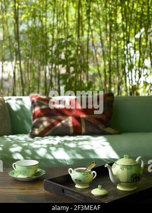 Getupftes Sonnenlicht im Outdoor-Sitzbereich mit china-Teekanne auf Holztisch mit Union Jack Kissen und Bambus Hintergrund in Wohnhaus, USA. Stockfoto