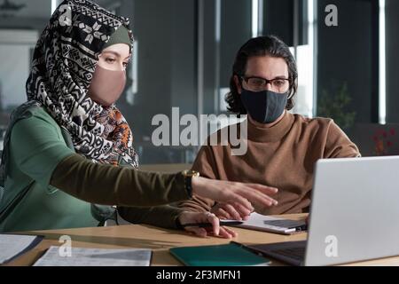 Junge muslimische Frau in Maske diskutiert Online-Präsentation mit ihr Partner während der Besprechung im Büro Stockfoto