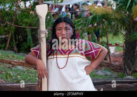 Ein Porträt einer jungen Maya-Frau mit Make-up und traditioneller Kleidung in ihrem Dorf in Mexiko. Tropische Vegetation und andere Menschen im Hintergrund. Stockfoto