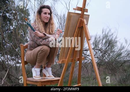 Blonde junge Frau Malerei in einem Feld mit Pinsel und Staffelei. Künstlerkonzept. Stockfoto