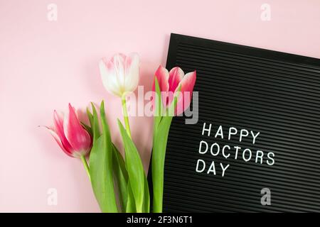 Schwarzer Briefbogen mit weißen Plastikbuchstaben mit Zitat Happy Doctor's day und einem Bündel rosa Tulpen auf rosa Hintergrund. National Doctors Day. Glücklicher Krankenschwester Tag. Flach lag Stockfoto