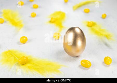 goldenes Ei auf weißem Betongrund mit gelben Federn und gelben Pralinen in Form eines Eies. Frohe Ostern Konzept. Weichfokus Stockfoto