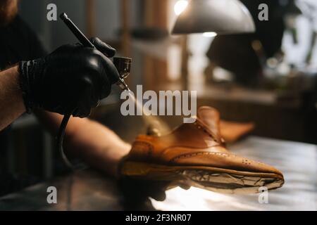 Nahaufnahme Hände von nicht erkennbaren Schuster tragen schwarze Handschuhe Sprühen Farbe von hellbraunen Lederschuhe. Stockfoto