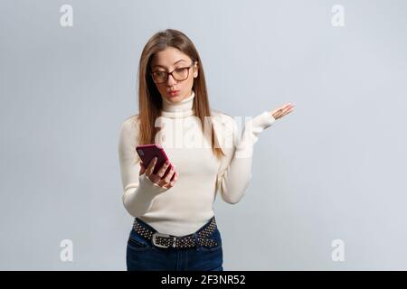 Ein schönes Mädchen in Brille schaut auf den Smartphone-Bildschirm in Überraschung. Eine junge braunhaarige Frau nutzt ein Mobiltelefon, um Nachrichten auszutauschen. Modern te Stockfoto