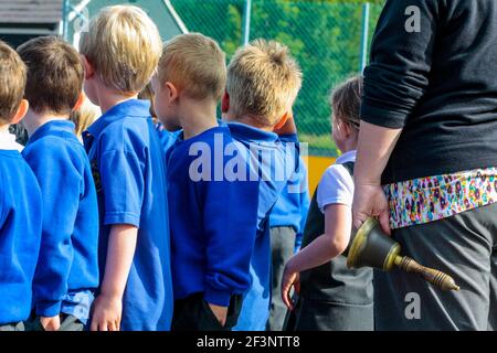 Gruppe von Grundschulkindern in Schuluniform, die nach einer Pause im Unterricht neben einem Lehrer, der Glocke hält, ihren Weg zurück in das Klassenzimmer machen. Stockfoto