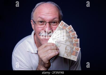 Weißer Erwachsener Mann lächelt an der Kamera und hält mehrere Brasilianer Echtgeldrechnungen Stockfoto