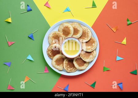 Teller mit kleinen runden Pfannkuchen mit bunten Party Topper Flaggen auf Split dreifarbig geschichteten Papier Hintergrund. Draufsicht, kräftige, lebendige Farben, Orange Stockfoto