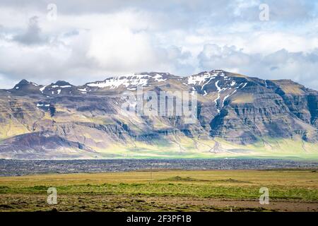 Landschaftsansicht bei Gerduberg Basaltsäulen in Snaefellsnes Island mit Grünes Gras am Sommertag und Wolken am Himmel und Schneebedeckter vulkanischer Berg Stockfoto