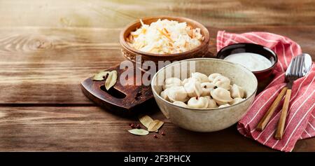 Traditionelle russische Pelmeni-Knödel mit Sauerrahm und Sauerkraut mit Sauerrahm auf Holzgrund. Stockfoto