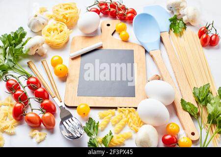 Kleine Tafel mit Kopierplatz umgeben von Zutaten zum Kochen verschiedene Arten von Pasta, Gewürze, Utensilien, gesunde rohe Gemüse Scattere Stockfoto