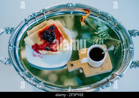 Blick aus dem hohen Winkel auf die silberne Frühstücksplatte mit Pfannkuchen und Marmelade und Kaffee, mit einer orangefarbenen Blume auf der Seite Stockfoto