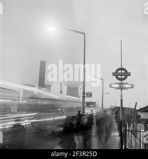 HARLESDEN, Großraum London. Eine Straßenszene mit Verkehr und Fußgängern, die nachts an einer Bushaltestelle an einem Schild der Londoner U-Bahn-Station in Harlesden vorbeifahren, Stockfoto