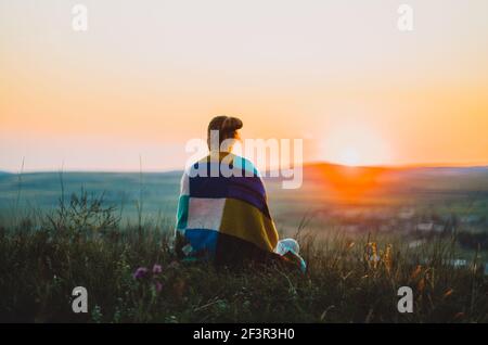 Rückansicht einer jungen Frau, eingewickelt in eine bunte Strickdecke, sitzend auf einem Hügel bei Sonnenuntergang, meditierend, kontemplierend Stockfoto