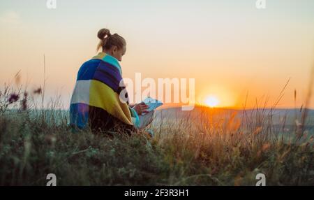 Rückansicht einer jungen Frau, eingewickelt in eine bunte Strickdecke, sitzend auf einem Hügel bei Sonnenuntergang, lesend, meditierend, kontemplierend Stockfoto