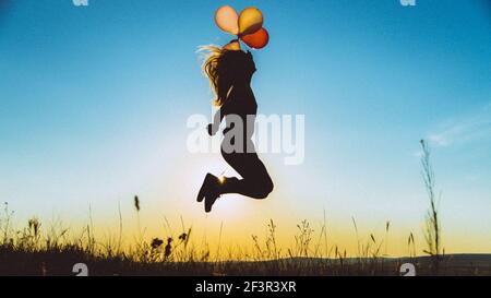 Goldene blaue Silhouette einer jungen Frau springen mit Ballons auf einem Hügel von Sonnenuntergang goldenes Licht, Konzept der Freiheit und Unachtsamkeit hinterleuchtet Stockfoto