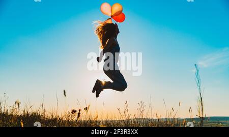 Goldene blaue Silhouette einer jungen Frau springen mit Ballons auf einem Hügel von Sonnenuntergang goldenes Licht, Konzept der Freiheit und Unachtsamkeit hinterleuchtet Stockfoto