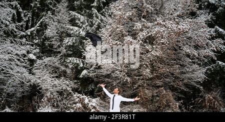 Fröhlicher junger Geschäftsmann, der in der verschneiten Winternatur steht und den Erfolg feiert, indem er seine Jacke hoch in die Luft wirft. Stockfoto