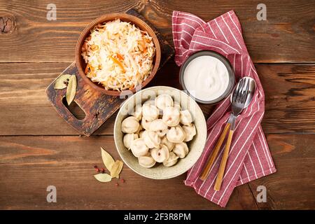 Traditionelle russische Pelmeni-Knödel mit Sauerrahm und Sauerkraut mit Sauerrahm auf Holzgrund. Draufsicht, flach liegend. Stockfoto