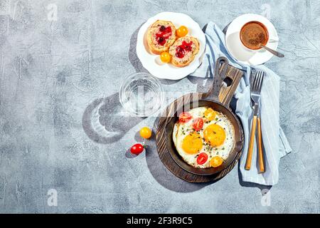 Spiegeleier. Spiegeleier aus zwei Eiern in gusseiserner Pfanne mit Kirschtomaten und Microgreens, Toast und Tasse Kaffee. Konzept für ein sonniges Frühstück am Morgen. Stockfoto