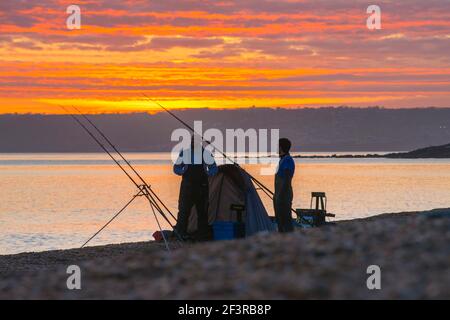 Seatown, Dorset, Großbritannien. 17th. März 2021. Wetter in Großbritannien. Zwei Fischer, die den spektakulären Sonnenuntergang vom Strand aus betrachten, während sich die Wolken beim Sonnenuntergang in Seatown in Dorset rot färben, während der Himmel nach einem bewölkten Nachmittag während der Covid-19-Sperre aufklärt. Bild: Graham Hunt/Alamy Live News Stockfoto