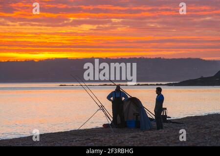 Seatown, Dorset, Großbritannien. 17th. März 2021. Wetter in Großbritannien. Zwei Fischer, die den spektakulären Sonnenuntergang vom Strand aus betrachten, während sich die Wolken beim Sonnenuntergang in Seatown in Dorset rot färben, während der Himmel nach einem bewölkten Nachmittag während der Covid-19-Sperre aufklärt. Bild: Graham Hunt/Alamy Live News Stockfoto