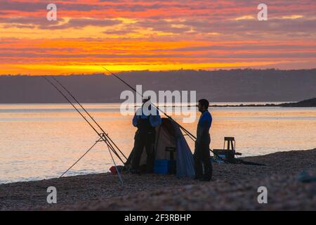 Seatown, Dorset, Großbritannien. 17th. März 2021. Wetter in Großbritannien. Zwei Fischer, die den spektakulären Sonnenuntergang vom Strand aus betrachten, während sich die Wolken beim Sonnenuntergang in Seatown in Dorset rot färben, während der Himmel nach einem bewölkten Nachmittag während der Covid-19-Sperre aufklärt. Bild: Graham Hunt/Alamy Live News Stockfoto