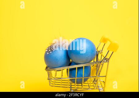 Drei große blaue Eier in einem Supermarkt-Trolley auf einem hellgelben Hintergrund. Ostershopping-Konzept Stockfoto