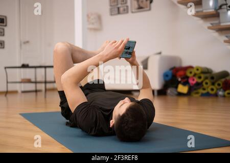 Fit Mann ruht nach harter Fitness und SMS mit Freunden auf dem Handy. Ruhezeit. Mann liegt auf schwarzer Yogamatte und Messaging per Smartphone. Stockfoto