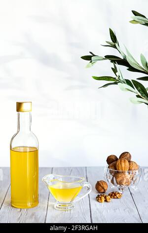 Walnussöl und ein Bündel Nüsse auf einem weißen Holztisch. Modernes Stillleben mit Ölflasche und Glasschale mit Walnüssen, Olivenzweig und Kopierraum. Stockfoto