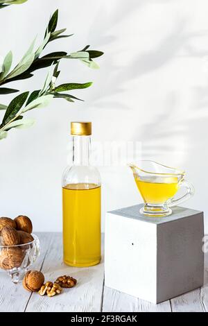 Walnussöl und ein Bündel Nüsse auf einem weißen Holztisch. Modernes Stillleben mit Ölflasche und Glasschale mit Walnüssen, Olivenzweig und Kopierraum. Stockfoto