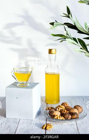 Walnussöl und ein Bündel Nüsse auf einem weißen Holztisch. Modernes Stillleben mit Ölflasche und Glasschale mit Walnüssen, Olivenzweig und Kopierraum. Stockfoto