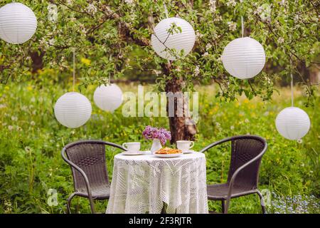 Niedliches Arrangement mit Gartenmöbeln, zwei Teetassen und Kuchen auf dem Tisch. Chinesische Papierlaternen, die zur Dekoration vom blühenden Apfelbaum hängen. Stockfoto