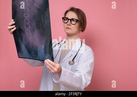 Ärztin in einer Brille, die auf eine Röntgenaufnahme schaut. Studio auf rosa Wand aufgenommen. Stockfoto