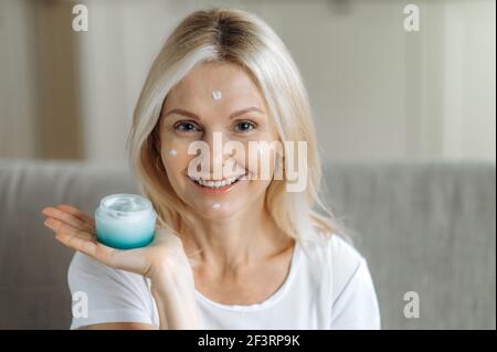Kaukasische blonde reife Frau lächelt und schaut auf die Kamera. Erstaunliche Erwachsene Dame hält eine Creme in der Hand, feuchtigkeitsspendende Gesichtscreme in ihrem Gesicht, trägt Tropfen der Creme auf ihr Gesicht, Hautpflege Stockfoto
