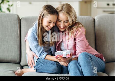 Mutter und Tochter umarmen sich gegenseitig. Eine blonde kaukasische Mutter und ihr Vorschulmädchen sitzen auf dem Sofa im Wohnzimmer und schauen auf die Geschenkbox, wunderschön dekoriert mit einer Schleife, lächelnd Stockfoto